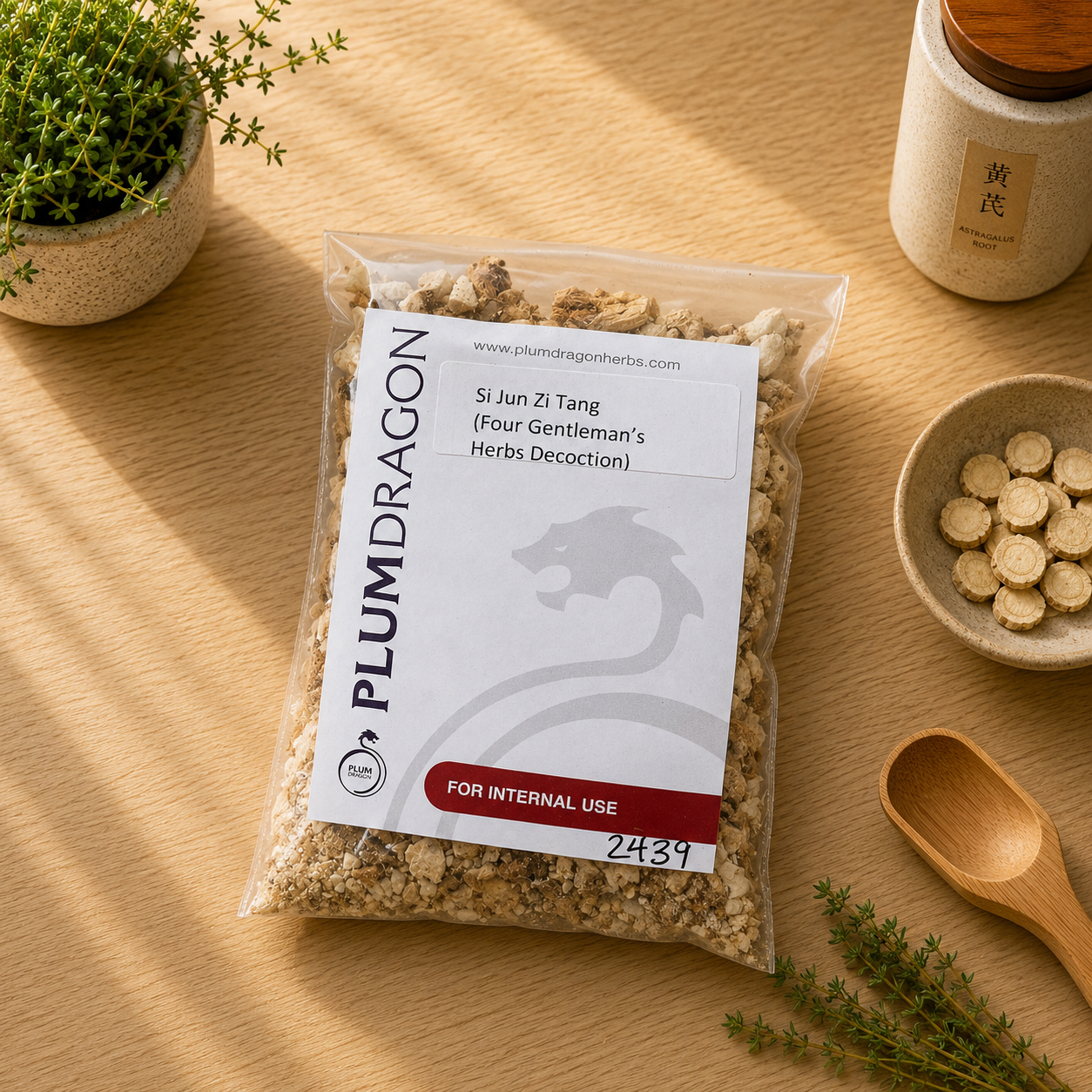 A clear plastic bag of Plum Dragon Si Jun Zi Tang herbal decoction sits on a light wood tabletop with small herb plants, a spoon, and a small bowl of dried tablets nearby. The package has a white label with black and red text and branding.