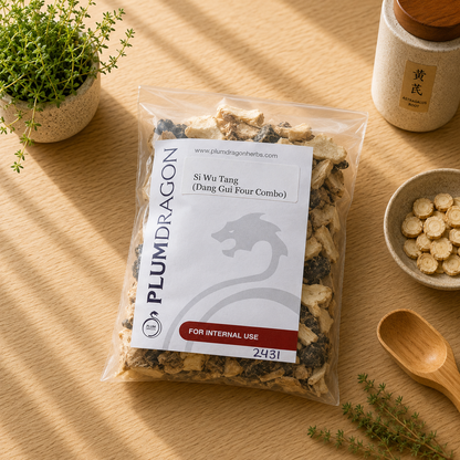 A clear plastic bag of Plum Dragon's Si Wu Tang herbal powder sits on a light wood tabletop, surrounded by small plants, a ceramic jar, a bowl of sliced herbs, and a wooden spoon. The label reads 'Si Wu Tang (Dang Gui Four Combo)' and 'FOR INTERNAL USE'.