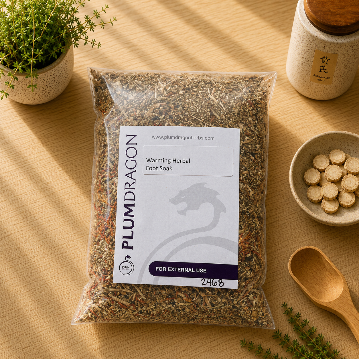 A clear plastic bag of Plum Dragon's Warming Herbal Foot Soak sits on a light wood table, surrounded by small potted plants, a ceramic jar, a bowl of round herbal tablets, and wooden spoons. 